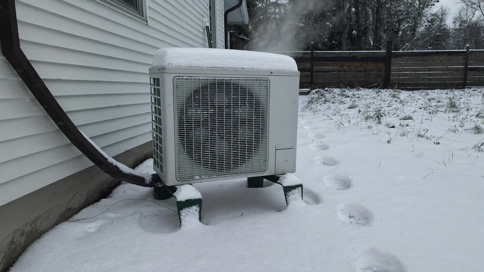 Cold-climate heat pump outdoor unit on a stand surrounded by snow beside a Massachusetts house in February